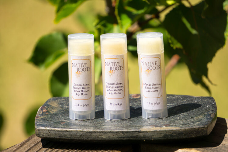 Dervaes+Institute+-+lipbalm Three Native Roots lip balm tubes displayed on a stone surface, featuring flavors: Lemon-Lime, Vanilla Bean, and Mango Butter. Each tube contains moisturizing mango butter and shea butter, ideal for lip care. Green foliage is blurred in the background, emphasizing the natural ingredients.