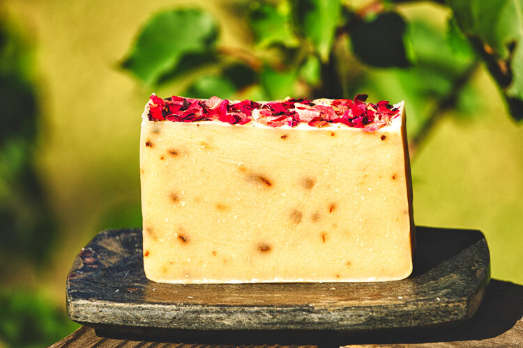 Dervaes+Institute+-+soap Natural handmade soap with rose petals, displayed on a wooden plate against a blurred green background. Ideal for skincare enthusiasts seeking organic and aesthetically pleasing bath products.
