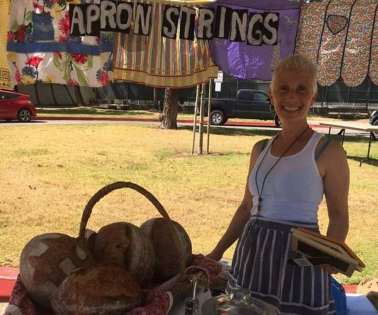 Woman smiling at a market stall under a colorful canopy, showcasing artisanal bread and handmade goods, with a sign reading "Apron Strings" in the background.