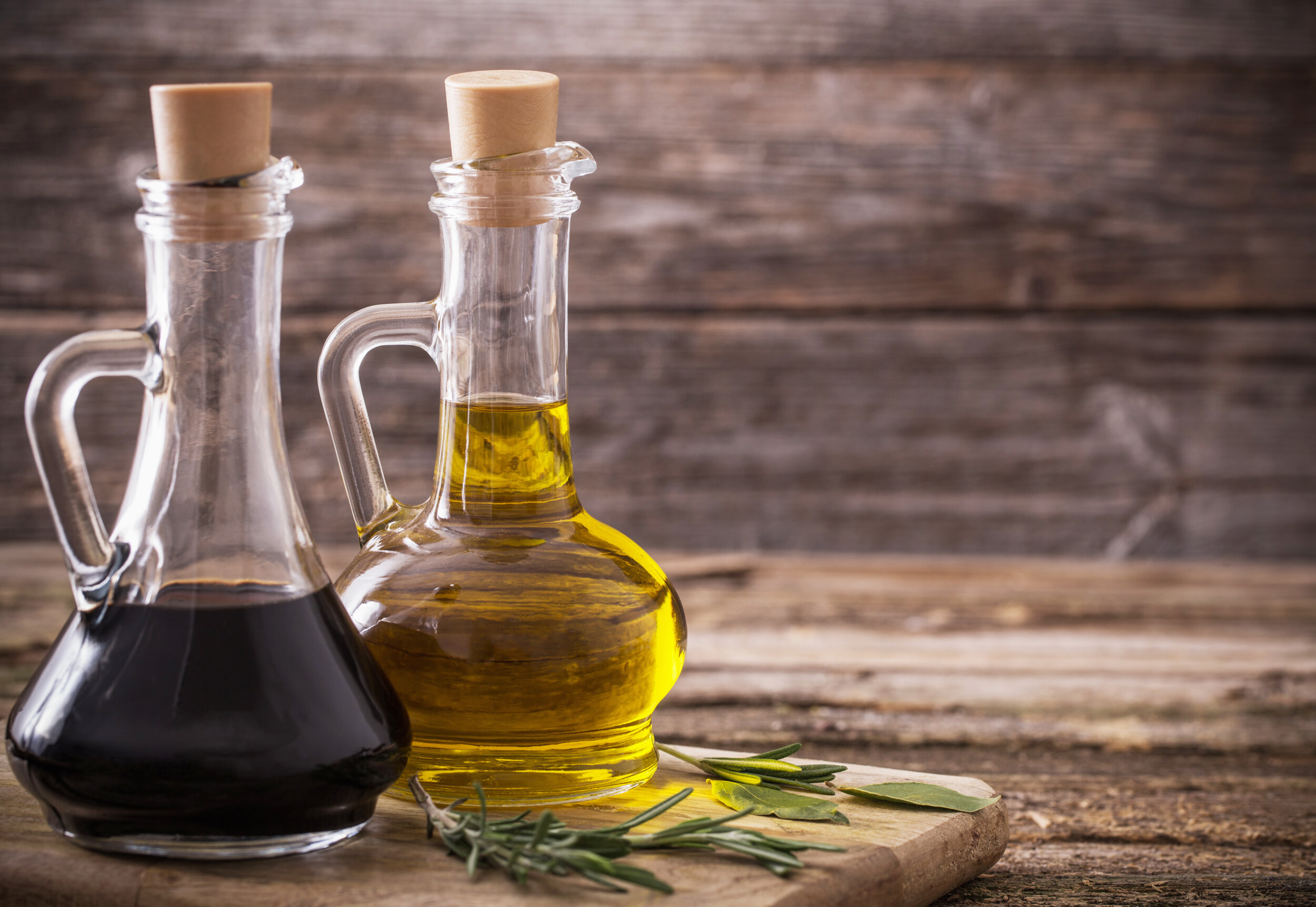 olive oil and balsamic vinegar on a wooden background Two glass bottles containing olive oil and balsamic vinegar, placed on a wooden cutting board with fresh herbs, set against a rustic wooden background. Ideal for culinary and cooking-related content.
